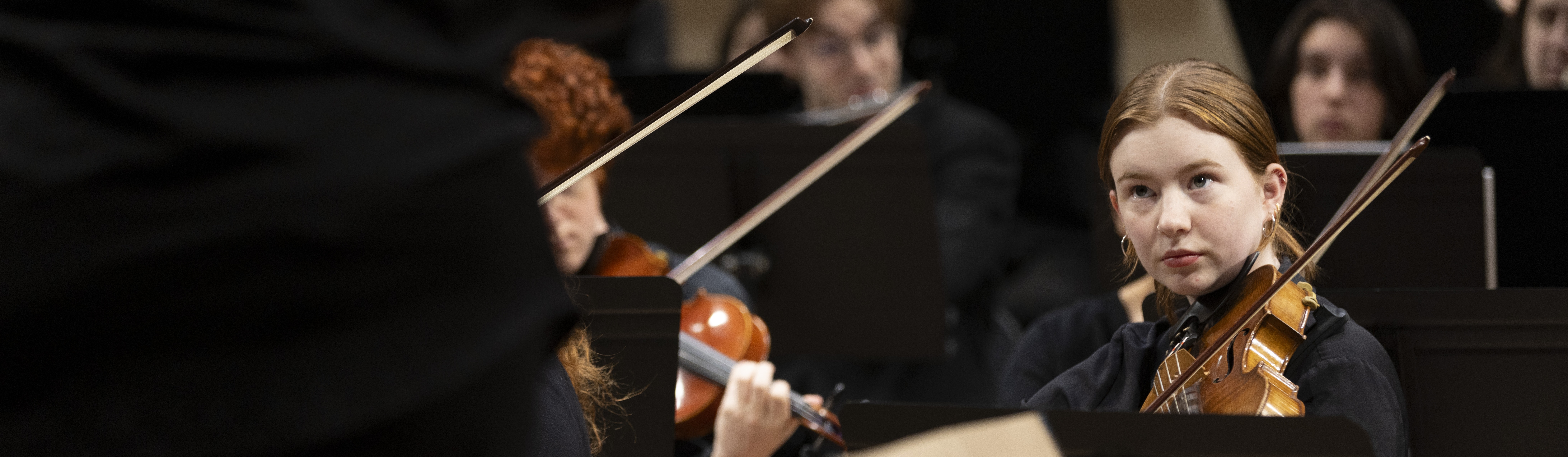 Close up of a student's hands as they play the C-flute with a conductor in the background.
