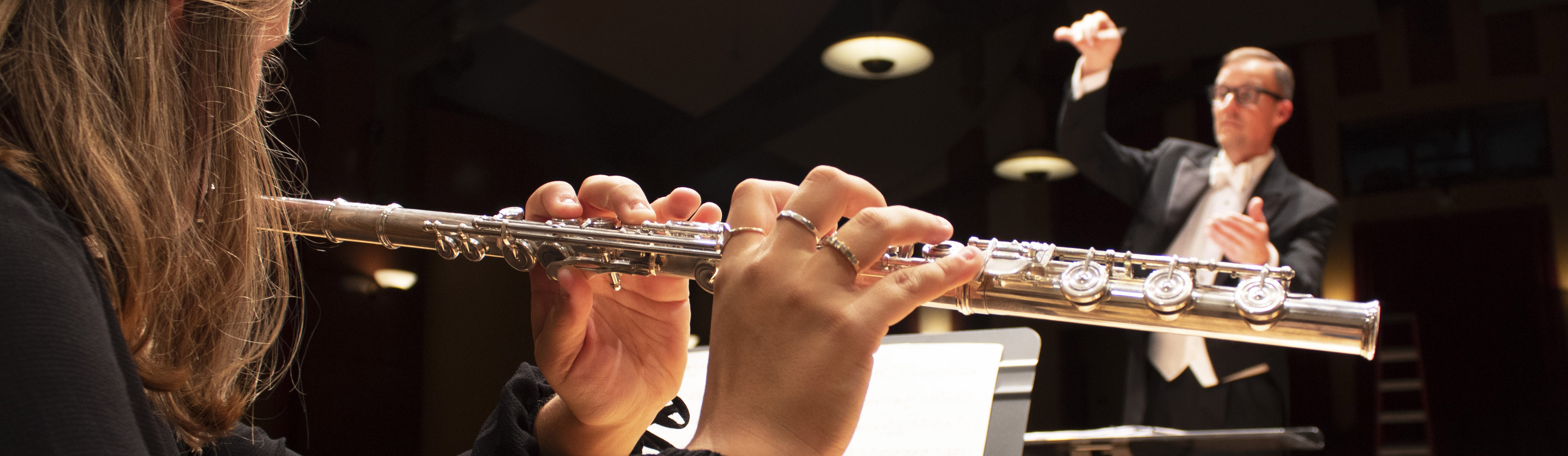Close up of a student's hands as they play the C-flute with a conductor in the background.